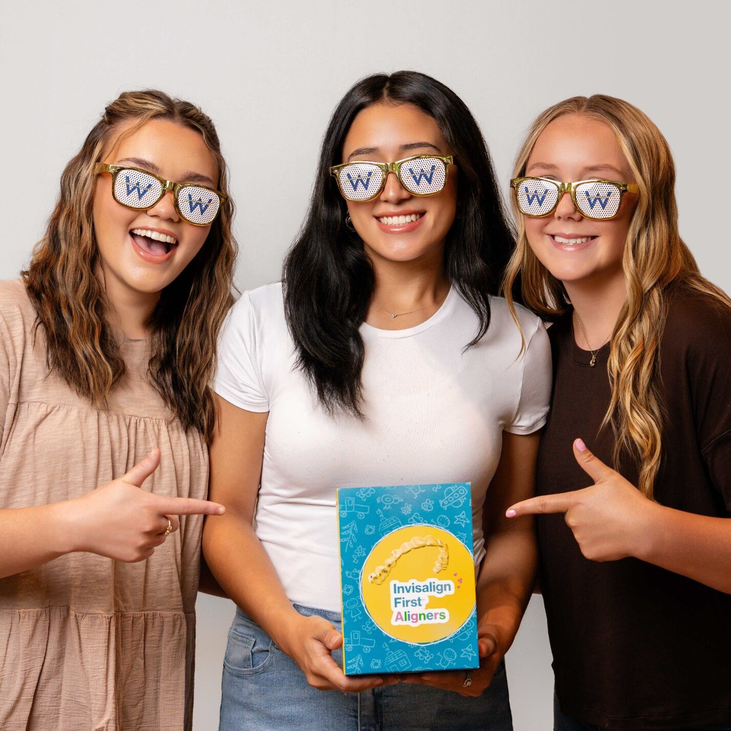 Three smiling young women wearing fun glasses with the letter "W" pose together, holding a box labeled "Invisalign First Aligners," promoting a friendly and welcoming atmosphere associated with Webb Orthodontics.
